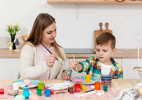 Woman and child painting Easter eggs at a table with art supplies.