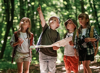 Children exploring a forest holding a map, wearing casual summer clothes.